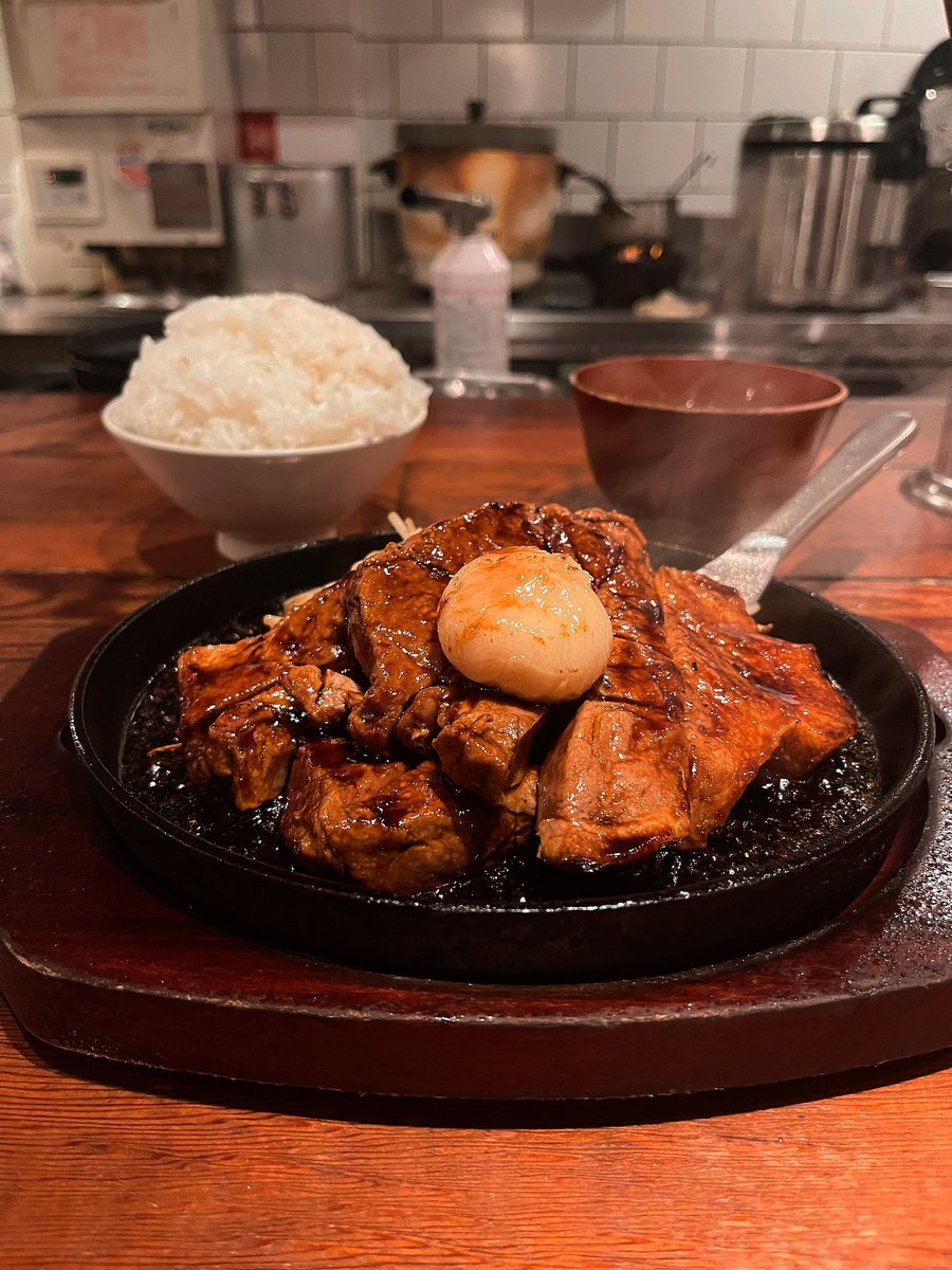 selfcomestomine's tweet photo. トンテキを食べながら思うことはひとつだ。人類って、こういう「高品質な食事」によって脳をデカくしてきた動物なんだよなーと。

脳というやつは、とにかく燃費が悪い。体重のわずか2%しかないくせに、安静時でも体全体のエネルギーの20〜25%を平然と消費する。

つまり、超高性能なCPUを四六時中フル稼働させているようなものだ。

問題は、その莫大な電気代をどう払うかである。その明らかな答えの一つは、高品質な食事だろう。

そして、ここから人類進化の“究極の家計簿”が始まる。

質の低い植物（葉や茎など）を主食にすると、大量に食べなければならず、長い消化管が必要になる。消化そのものにも、多くのエネルギーがかかる。

だが肉のような高品質な食事なら、少ない量でも十分なエネルギーが得られる。つまり、大きくて長い消化器官を一生維持する理由が薄くなるのだ。

1995年、人類学者レスリー・アイエロとピーター・ウィーラーは、有名な「高価な組織仮説」を提唱した。Aiello & Wheeler (1995). 

簡単に言えば、

「脳が大きくなったぶん、腸が小さくなった」

という進化のトレードオフ仮説である。

実際、ヒトの消化器官は霊長類として見ると異様にミニサイズだ。

まず胃。ヒトの胃の表面積は、同じ体重の典型的な哺乳類の3分の1にも満たず、ほかの97%の霊長類よりも小さい。

さらに大腸。同じ体重の霊長類なら期待されるサイズの60%以下しかない。大腸は植物の食物繊維を腸内細菌に発酵させ、脂肪酸というエネルギー源を作り出す巨大な発酵タンクだ。つまりヒトは、大型類人猿ほど植物からエネルギーを搾り取る能力を持っていない。

そして消化器官全体で見ても、ヒトの胃腸は同じ体格の霊長類から予測される重さの約60%しかない。

つまり、人類は本当に「腸を小さくした動物」なのである。

では、浮いたエネルギーはどこへ消えたのか？

答えはもちろん、脳だ。

進化というのは倹約家なので、オカンが家計の出費をケチケチ見直すように、まず維持費のかかる巨大な胃腸をダウンサイジング��る。

そして、そこで浮いた予算を、脳という電気食いマシンの開発費へ回した。

まとめると、人類進化ではこういうことが起きたらしい。

食事の質が上がる〜消化管が小さくなる〜維持コストが浮く〜浮いたエネルギーを脳へ投資する〜脳がさらに大きくなる

つまり人類とは、

腸を削って脳に課金した動物

なのである。
*R.Wrangham (2009). 

───そして、その課金システムを支えた重要な要素こそ、この目の前のトンテキみたいな「高品質な肉」だったわけだ。

📍トンテキ食堂なかむら （新宿三丁目）

#進化グルメ学