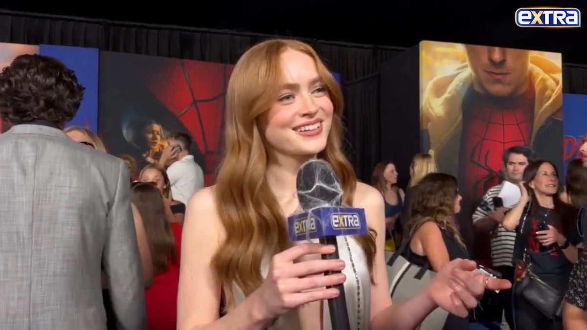 Actress Sadie Sink with long red hair smiles during an interview holding an "Extra" microphone at a Spider-Man premiere event.