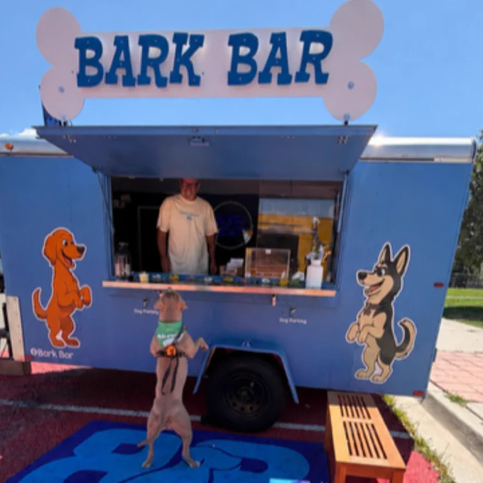 A small blue food truck named Bark Bar serves dog treats with cartoon dog illustrations on its sides and a dog standing on hind legs waiting at the counter.