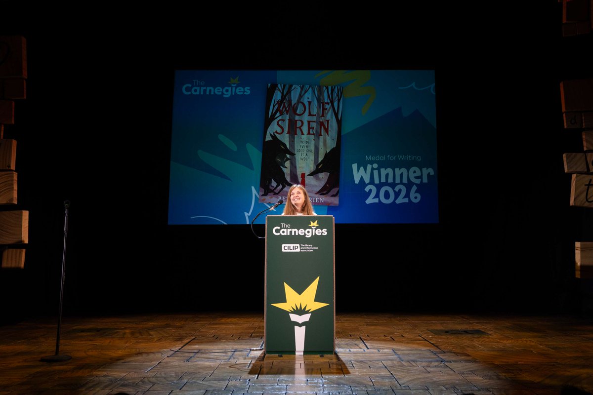Beth O'Brien stands at a podium on stage smiling after receving the 2026 Carnegie medal for writing with her book cover for Wolf Siren displayed behind her.