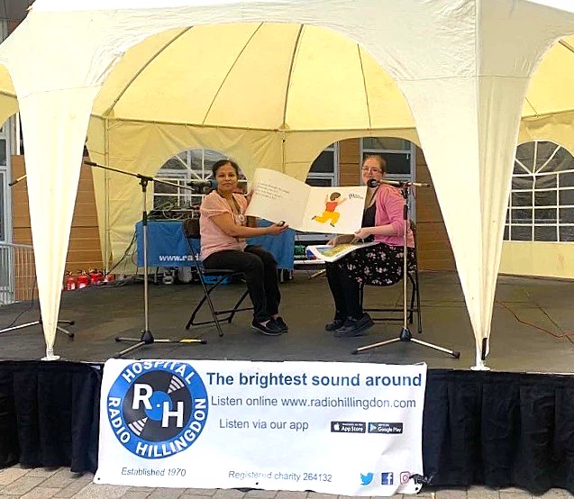 Photograph of 2 members of Hillingdon Libraries staff sat underneath a gazebo holding a large oversized children's book to be read to the crowd. Both are seated facing the camera.