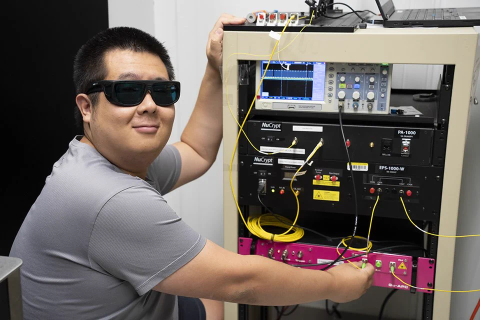 A person wearing sunglasses is seated next to electronic equipment, connecting yellow cables to a rack of devices. The rack contains various equipment including a device labeled "EPS-1000". There is a laptop on top of the rack, and some cables are visibly attached to it.