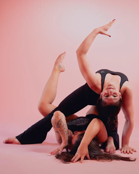 An image of Hannah & Morgan wearing black against a pink studio backdrop. The two make bold shapes with their body using contortion, Morgan lays on the floor with leg pointed upwards, whilst Hannah is leaning back and mirroring the shape of Morgan's leg with her arm.