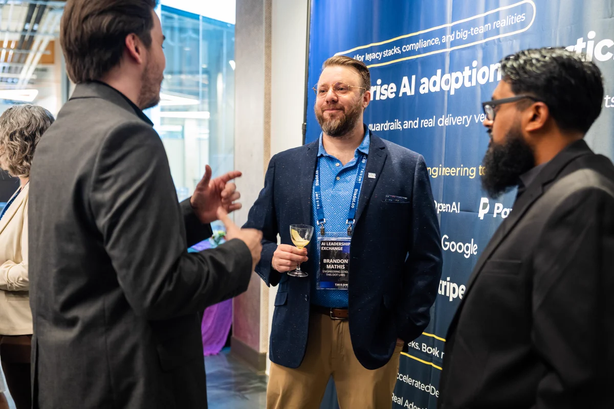 Three attendees network at a professional technology event in front of a This Dot Labs AI booth. The person in the center, wearing a conference badge and holding a glass, is engaged in conversation with two other attendees in business attire. The booth backdrop highlights enterprise AI adoption and features logos from companies including OpenAI, Google, Stripe, and PostgreSQL. The setting is a modern conference venue with additional attendees visible in the background.