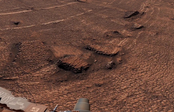 Reddish desert terrain textured with a pattern of small, hexagonal raised ridges. Part of the rover can be seen in the lower left. 