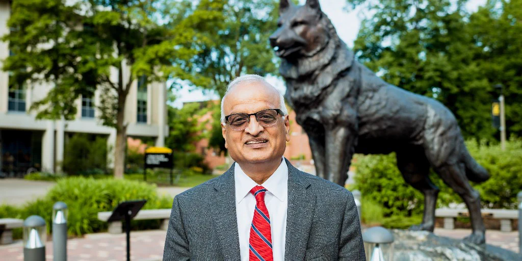 A photo of Sri Beldona, the new Michigan Tech Dean of the College of Business. He is wearing a gray suit and red and blue tie, and is posing in front of the Husky statue on Michigan Tech's campus.