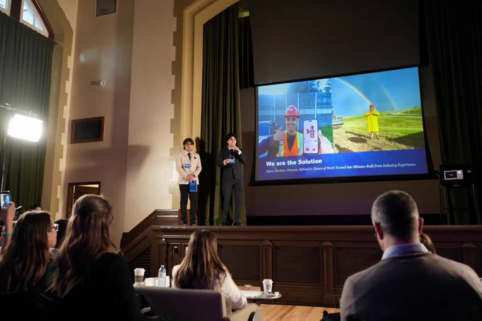Team BAETT is standing on a stage next to a large screen presenting a slideshow to an audience. The slide is titled "We are the Solution" and features images of a person in a hard hat and another holding a tablet, along with a wind turbine and a rainbow in the background. 