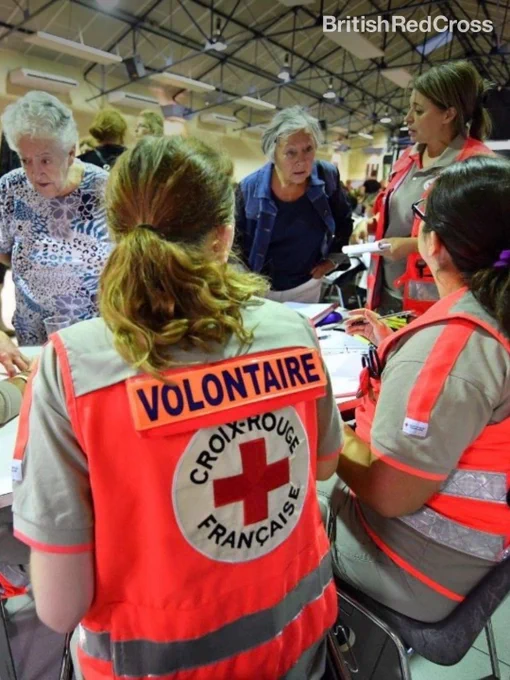 Photo of two volunteers from the French Red Cross wearing high vis vests. They are inside a building talking to two older women.