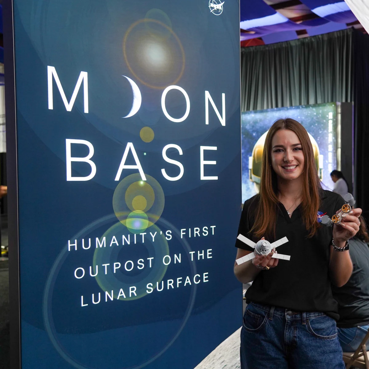 NASA Moon Base Program intern Macie Landon smiles while standing beside a tall Moon Base display at a NASA outreach event. The display reads "Moon Base" with the tagline, "Humanity's First Outpost on the Lunar Surface." Macie holds 3D-printed models of the Orion spacecraft and a lunar rover. Credit: NASA