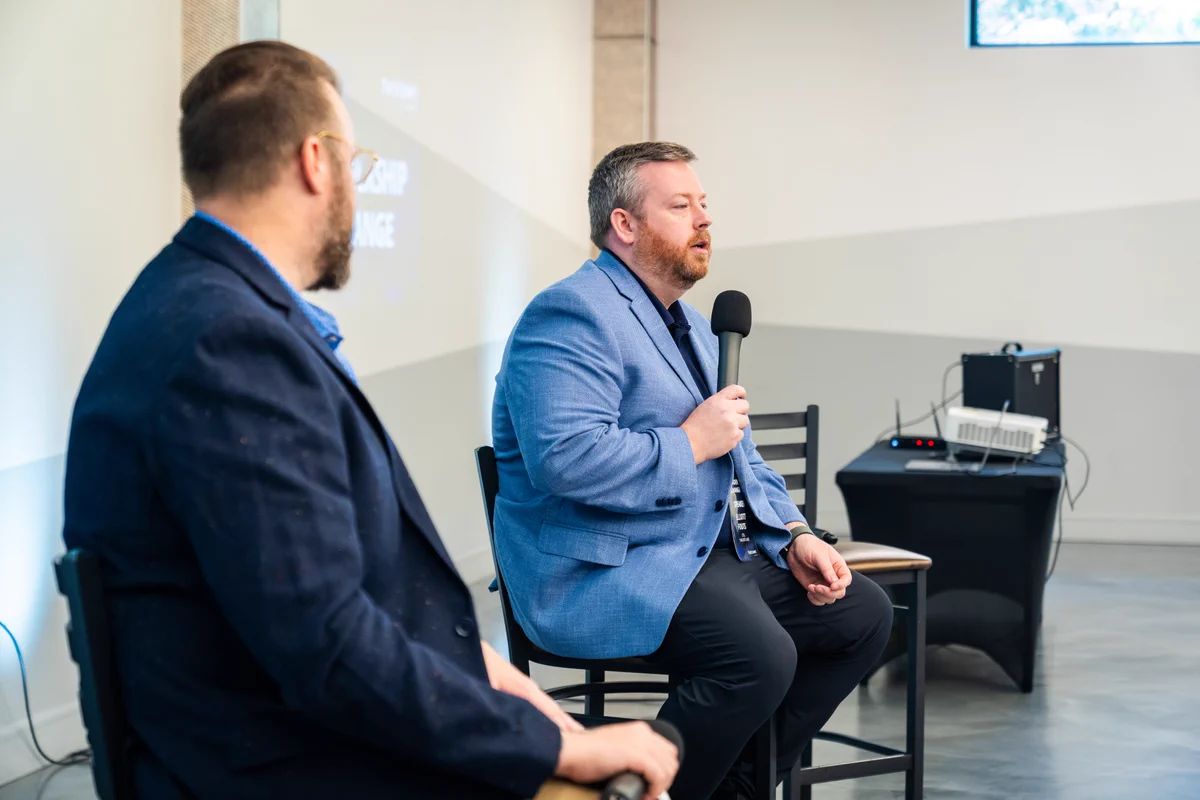 Two speakers participate in a seated panel discussion at an indoor event. One person in a light blue blazer speaks into a handheld microphone while the other listens from the foreground. A projector and presentation equipment are visible in the background, creating a professional conference setting.