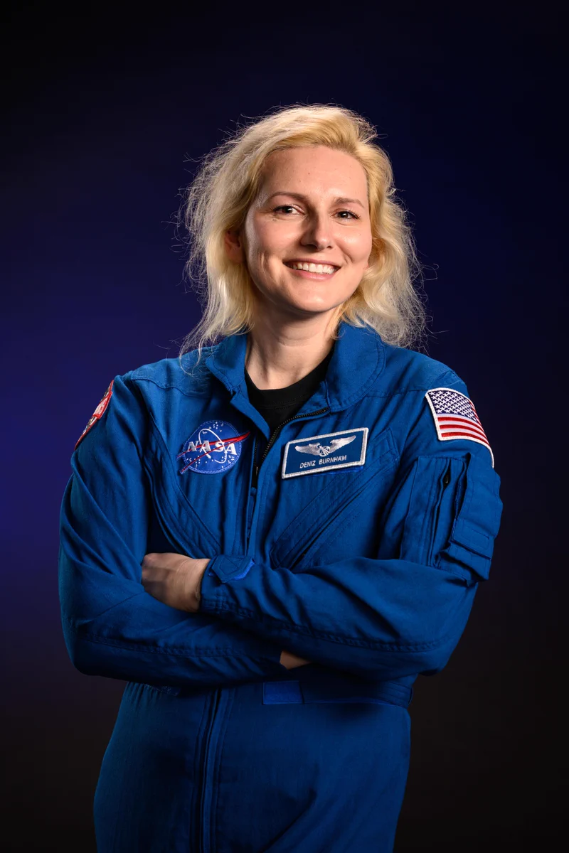 Astronaut Deniz Burnham, wearing her blue flight suit, stands and smiles at the camera with her arms crossed. The background behind her is a dark blue. Credit: NASA/Robert Markowitz
