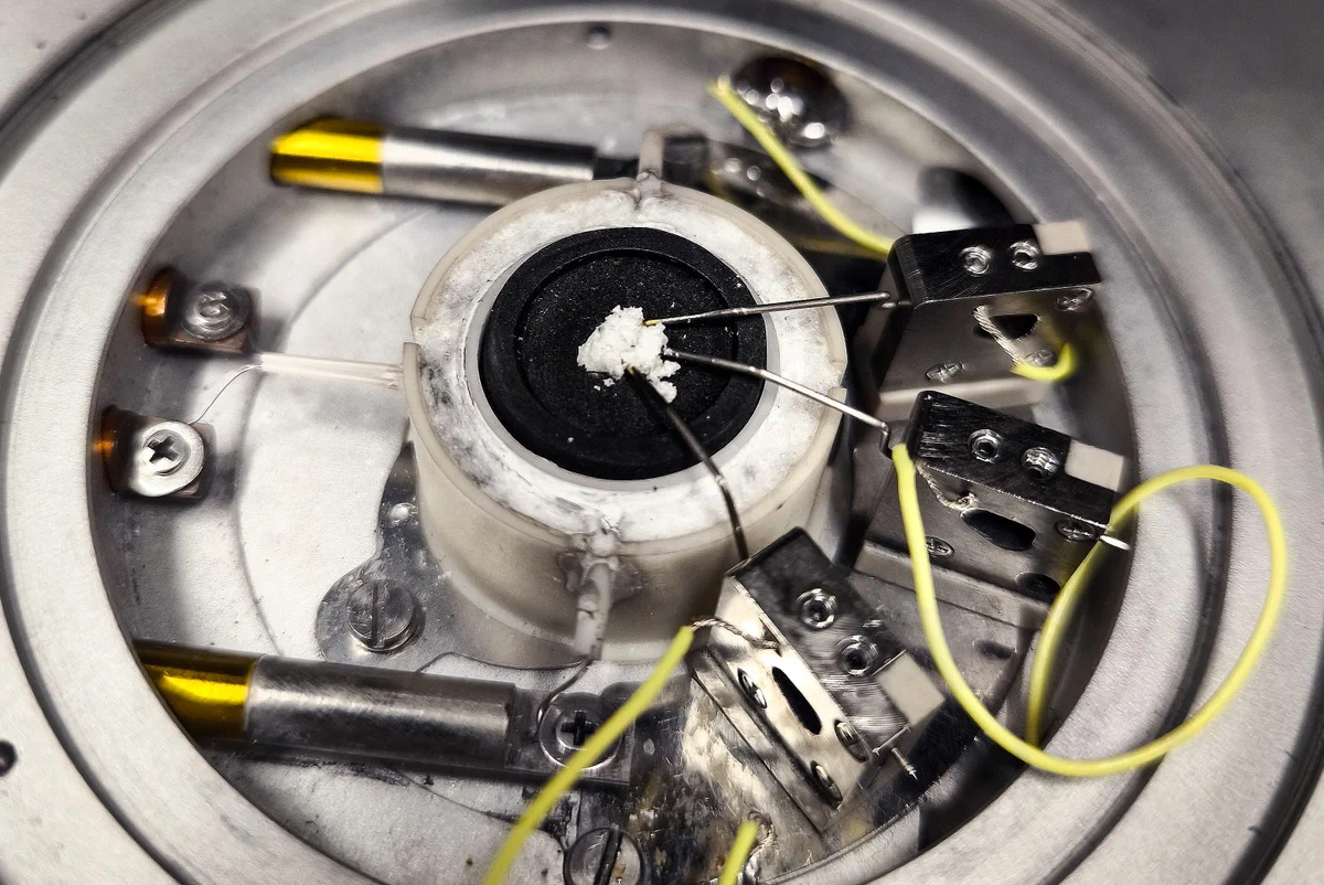 A scientific setup. At the center, a white clump of crystalline salt rests on a dark disc. Three fine yellow metallic wires extend from nearby metal blocks.
