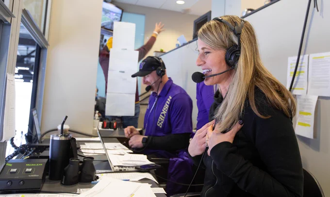 A smiling blonde woman in a headset and mic gestures excitedly, hands on her chest. A man in a purple 'HUSKIES' shirt and cap works on a laptop beside her. The desk has a 'Model 210' device, papers, and binoculars. Another person cheers in the background of this broadcast setting.
