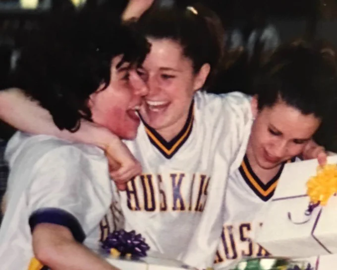 Three young women in white jerseys with purple and gold trim, featuring \"HUSKIES\" text, embrace and laugh heartily. Two women have arms around each other, smiling broadly. The woman on the right looks down at a white gift box with a yellow bow. Another box with a purple bow is visible.
