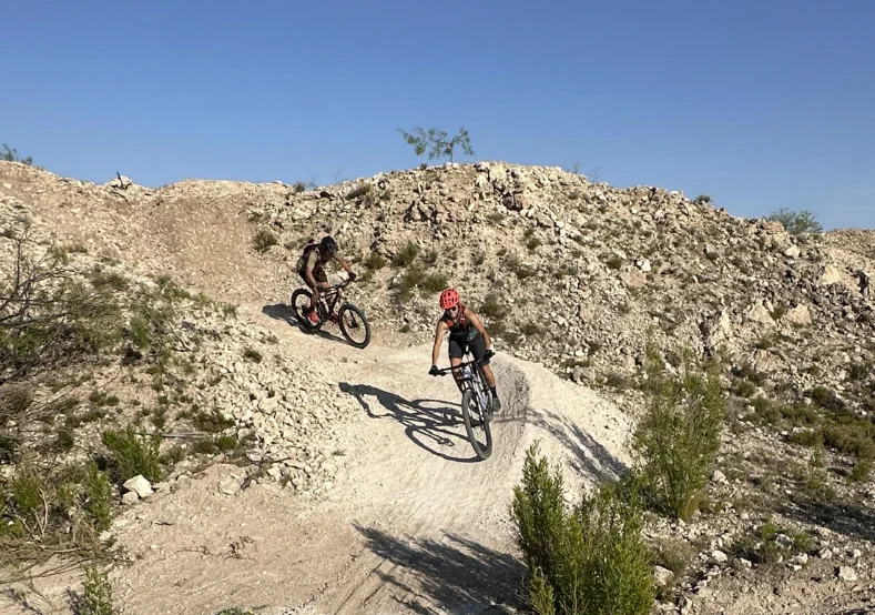 Two people mountain biking on a rocky trail under clear blue sky.