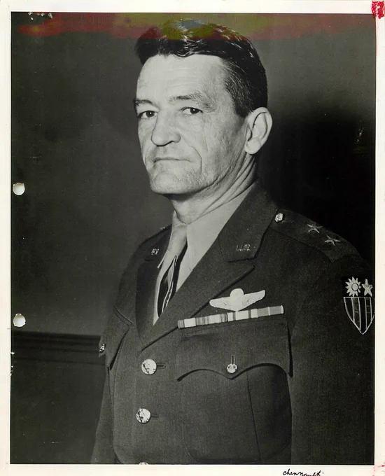 A person in a military uniform with various insignias stands facing forward. The uniform includes aviation wings, a double-star emblem on the shoulder, and a patch with a sun and three stars.