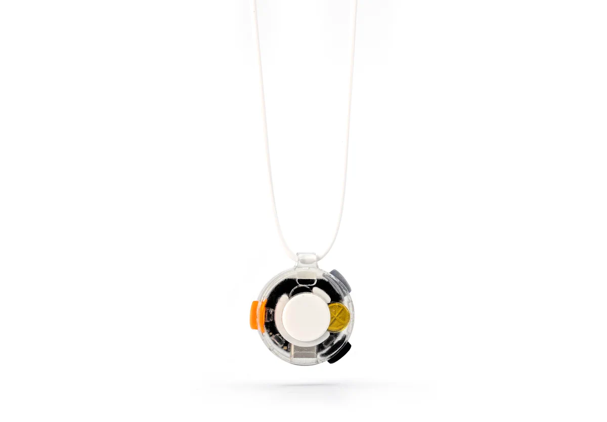 Vox, a small circular wearable device with a translucent housing, hanging from a thin white cord necklace against a white background. Internal components are visible through the clear enclosure, including a white circular disc at the center, a gold contact, and an orange side button.