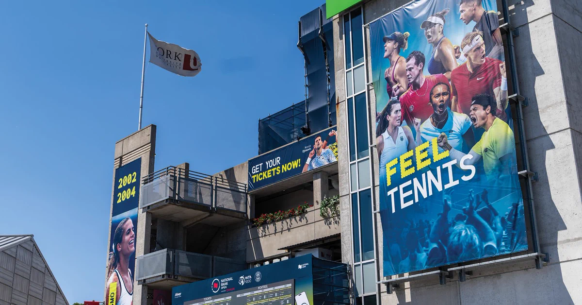 A sports stadium exterior with large banners featuring various tennis players. A prominent blue banner displays the text "Feel Tennis." Nearby, a flag is waving, and signage encourages ticket purchases. The years 2002 and 2004 are displayed on the building.