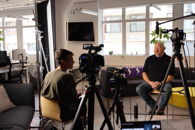 A person is seated in a well-lit office, speaking during an on-camera interview. Several cameras and lighting equipment are set up, with another individual appearing to monitor the recording process.