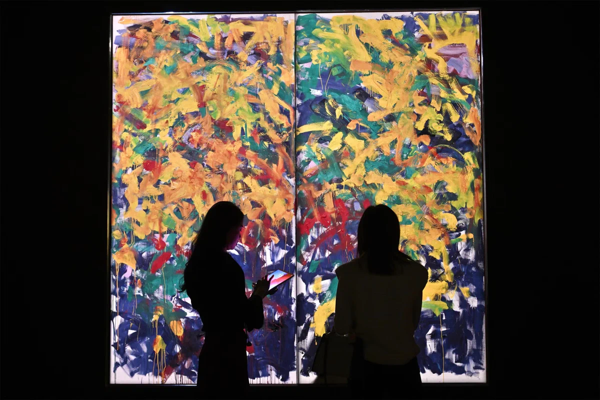 Two people stand in front of a large, colorful abstract painting, backlit to create a silhouette effect. The painting is vibrant, featuring splashes of yellow, blue, green, and red.