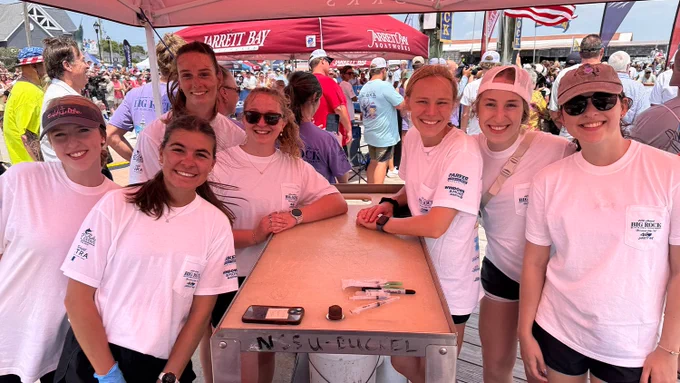 Group of seven young women in matching white t-shirts smiling around a wooden table at an outdoor event under red tents.