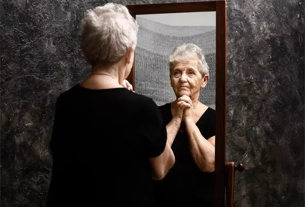 An older woman with short gray hair stands facing a mirror, holding her hands clasped together near her chin with a pensive expression.