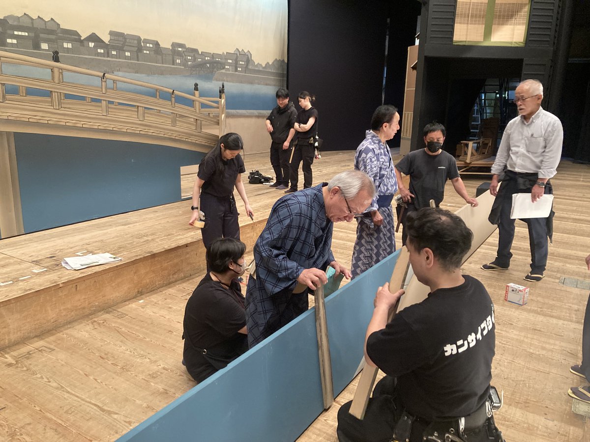 ono_hiroyuki's tweet photo. 【文楽『#まちの灯』振り返り６】
7月12日
この日は人形遣いの皆さんの稽古。前回の「場当たり」が全体のイメージをつかむシミュレーションだったとすれば、今日は実際に浄瑠璃の録音をかけながら最初から最後まで動きます。といっても、この日も主遣いの師匠方のみがいらっしゃって人形も持ちません。劇場の２階ロビーで、録音に合わせて演技をします。師匠方が人形になって動くのです。それにしても、人形がお上手な方は芝居がお上手。ご本人の芝居心・技術が、人形に伝わって増幅するのですね。
ほとんどの場面は師匠方が考えてこられた動きを１回あわせて確認。立ち位置の整理が必要な部分だけ、何度か返し稽古をしました。普段の古典の公演ではこのようなお稽古はありません。ツメ人形の皆さんに場面の気持ちをお伝えして立ち位置を決めたりしたのですが、それもこれまであまりなかったことだったらしいです。
懸案はラストシーンです。どう動くべきかについて勘十郎師・勘彌師と議論。あの奇蹟のクロースアップに込められたさまざまな意味と感情をどのように形にしたいかをお話しいたしました。映画だと一つのクロースアップで全てを表現するのですが、そこに奔流するあらゆる感情を分解分析して、人形の振り・表情の一つ一つ、浄瑠璃、三味線、照明、美術など総動員で文楽のラストシーンとして構築しなければなりません。難関です。
師匠方が事前に周到に準備してきてくださいましたのでスムーズに進みましたが、それでもこの日の稽古は合計６時間かかりました。

7月13日
この日は「道具調べ」。美術・大道具の確認です。新作文楽のために美術家が工夫を凝らした新しい美術に感動。１場面ずつ確認していきます。大仏は図面よりも少しかみてに変更するようお願い。幕を上げるタイミングや群衆の道具がガヤガヤする順番も決定。勘十郎師と蓑二郎師は川の杭の位置を決めて、大道具さんがその場で作ります。実際に技芸員が歩いてみて、「ここに平台が一段欲しいわ」となればテキパキと大道具さんが作業をします。大道具だけで��く、花籠を置く台の位置、花の色や本数などをどんどん決めていきます。小道具さんは花びらが落ちる仕掛けを作ってきてくれました。
私も神聖な国立文楽劇場の舞台にはじめてあがらせていただき緊張しました。（さすがに記念写真を撮ることは憚られました）

7月14日
この日のお稽古は、「立稽古」。「たて稽古」と読みます。舞台をやってる人なら、「実際に動いてみる、立ち稽古かな」と思ってしまいますが、文楽の世界では太夫と三味線のリハのことと言います。あくまで太夫と三味線の皆さんの通し稽古なので、舞台には幕をつり、人形遣いの皆さんは幕の後ろで音を聞きながら動いてみるか、楽屋に聴こえてくる音を聴いてシミュレーション稽古をするそうです。この日のリハは録音し、明日の稽古で使います。
改めて通して聞きましたが、素晴らしすぎます。字幕を出す人も一緒にお稽古です。
この時点でラストシーンの演出候補は「引き幕」か���緞帳」で終わるか、「ラストの三味線の長さをどうするか」など４パターンの候補があり、その分も録音しました。

7月15日
この日の稽古は「居処調べ」。前日の立稽古で録音した義太夫を流しながら、実際の舞台美術の中で人形遣いが演技をします。
吉矢さんから稽古冒頭に何か一言話しますか？と言われ、一言ご挨拶というのもおこがましいように感じましたが、改めて決意を述べさせていただこうと思い、「ユージーン・チャップリンさんから先ほど飛行機に乗ったという連絡をいただきました。とても楽しみにしておられます。歴史的な舞台、どうか成功しますようお願いします！」と申し上げました。その時、技芸員さんから拍手が起きて感激。この道数十年の師匠方がこれまでの経験と芸、何よりすごい情熱を込めて全身全霊で新作に取り組んでおられる。みんなが成功に向けて一丸となっているのを感じて、稽古が始まりました。
さてこの日もまだ人形は持ちません（正確には大仏のシーンだけ勘十郎師は人形をお持ちになり、確認なさっていました）。主遣いが実際に動きながら確認・修正。左と足遣いの皆さんは舞台脇に��ばりついて主遣いがどう動くかを見て、ご自分の動きを研究なさっています。いよいよ形になってきました。相撲シーンは爆笑もの。玉助さんのご提案で大文字屋のセットのしもてにあった格子を、お客様��ら座敷の中がよく見えるように、その場で外してもらいました。
前日まで４パターンの中で迷っていたラストは引き幕に決定し、照明のタイミング、幕を引くタイミングもばっちり決まりました！
#新作 #文楽 #チャップリン #国立文楽劇場 #街の灯

写真は道具調べ、立稽古、居処調べの様子。