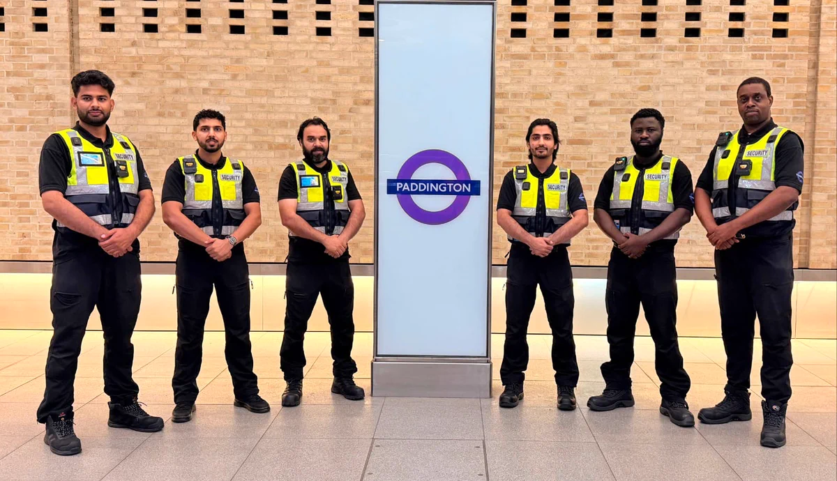 Six security guards, men of diverse backgrounds, stand uniformly in black clothing and bright yellow 'SECURITY' vests, hands clasped. They are positioned in front of a tall white sign with the purple Elizabeth line 'PADDINGTON' logo. A light brick wall is in the background.