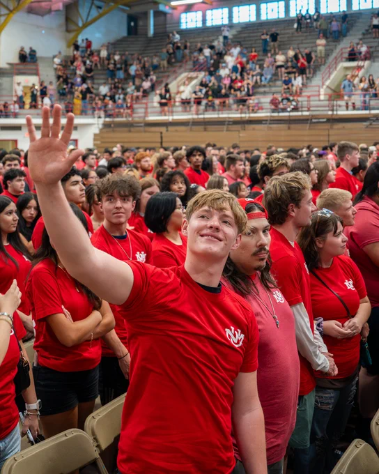 A large group of students wearing matching UNM shirts stands inside a gymnasium full of spectators in bleachers. One individual in the foreground is smiling and waving, while others around are looking in various directions.