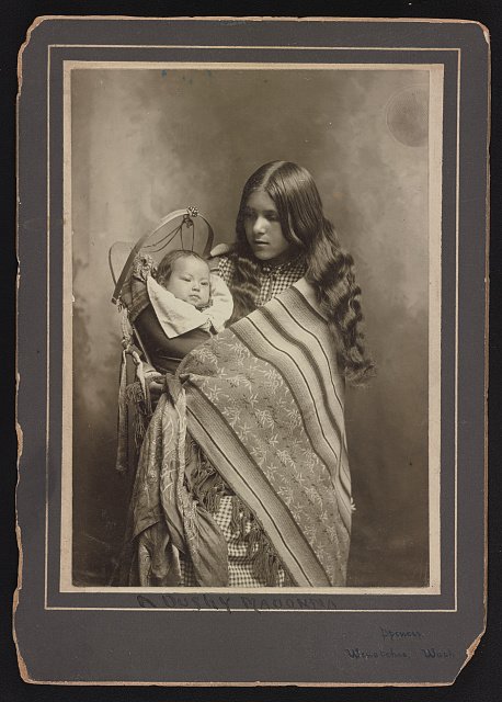 archeohistories's tweet photo. In 1902, a Plateau Indigenous woman carrying her baby in a cradleboard was doing far more than caring for a child. She was carrying knowledge that had passed through generations of women at a moment when the world around them was being radically altered.

Across the Columbia Plateau, women were central to family and economic life. They gathered roots and berries, processed and preserved food, made clothing, baskets and woven bags, and developed highly specialized artistic traditions. Knowledge of particular gathering places could pass through families, including from mothers to daughters.

Their craftsmanship could be remarkably complex. An early twentieth-century collector visiting Spokane communities encountered an elderly woman who was among the few people he found who still possessed knowledge of a difficult double-weaving technique. Such skills were not simply decorative. They represented generations of observation, teaching and memory.

The cradleboard belonged to this working world. It kept an infant close while allowing a woman's hands to remain free for travel, gathering and other responsibilities. Motherhood was not separated from daily work. Babies moved through the landscape with their mothers.

But in 1902, that landscape was changing.

The Spokane Reservation had been established in 1881, restricting Spokane people to a fraction of the territory their ancestors had traditionally used. Federal assimilation policies pushed the disruption further. Under the Dawes Act, communally held Native lands were divided into individual allotments, a policy designed in part to dismantle traditional tribal systems. Allotment of the Spokane Reservation began in 1902 and continued through 1908.

For women whose knowledge depended upon access to particular root beds, berry grounds, waterways and seasonal gathering places, losing land could also mean losing access to the physical geography of cultural memory.

By then, traditional food systems were already under pressure. Salmon resources had declined, access to hunting and gathering territories had contracted, and valuable agricultural lands throughout the region had increasingly passed into non-Native hands.

Yet Indigenous women continued raising children, producing intricate beadwork and weaving, gathering food when access permitted, and transmitting knowledge.

A cradleboard from this period therefore tells a much larger story. Inside it was a new generation being carried through one of the most disruptive periods in Plateau history, surrounded by women working to preserve knowledge that federal policy was actively trying to replace.

#archaeohistories