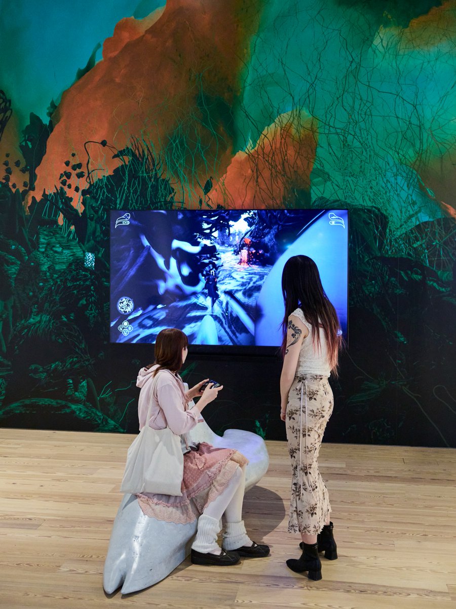 Two individuals are interacting with a digital installation at the Whitney. One is seated on a stylized white sculpture, holding a gaming controller, while the other stands nearby. A vibrant screen, surrounded by a colorful mural, displays a dynamic scene.
