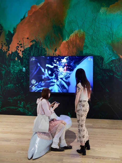 Two individuals are interacting with a digital installation at the Whitney. One is seated on a stylized white sculpture, holding a gaming controller, while the other stands nearby. A vibrant screen, surrounded by a colorful mural, displays a dynamic scene.