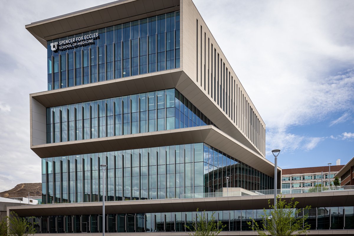 The image shows the Spencer Fox Eccles School of Medicine building, a modern multi-story structure with large glass windows and angular design features, set against a cloudy sky.