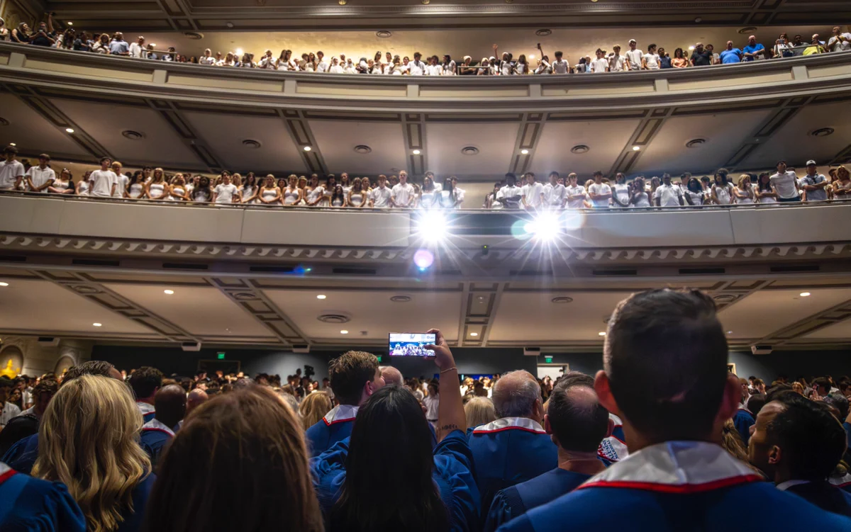 A large crowd in graduation gowns faces a stage in an auditorium. Bright lights shine toward the audience. Additional spectators fill the upper balcony wearing white, looking down toward the crowd.