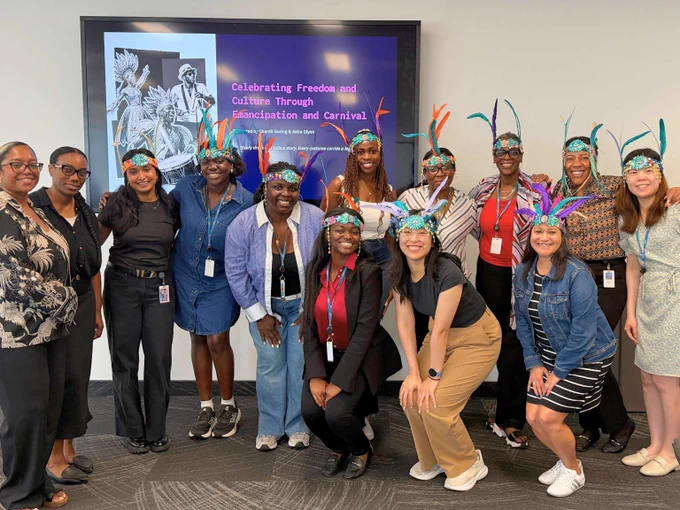 A diverse group of coworkers wearing colorful feathered headbands posing in front of a screen about Emancipation and Carnival celebration.
