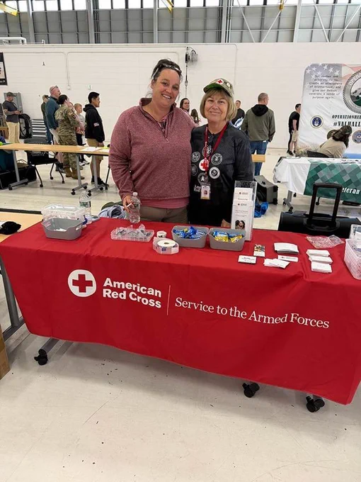A person wearing a red top and another wearing a cap stand behind a table covered with a red cloth displaying the American Red Cross logo and the words "Service to the Armed Forces." The table holds various items. Several people are visible in the background in a well-lit room.