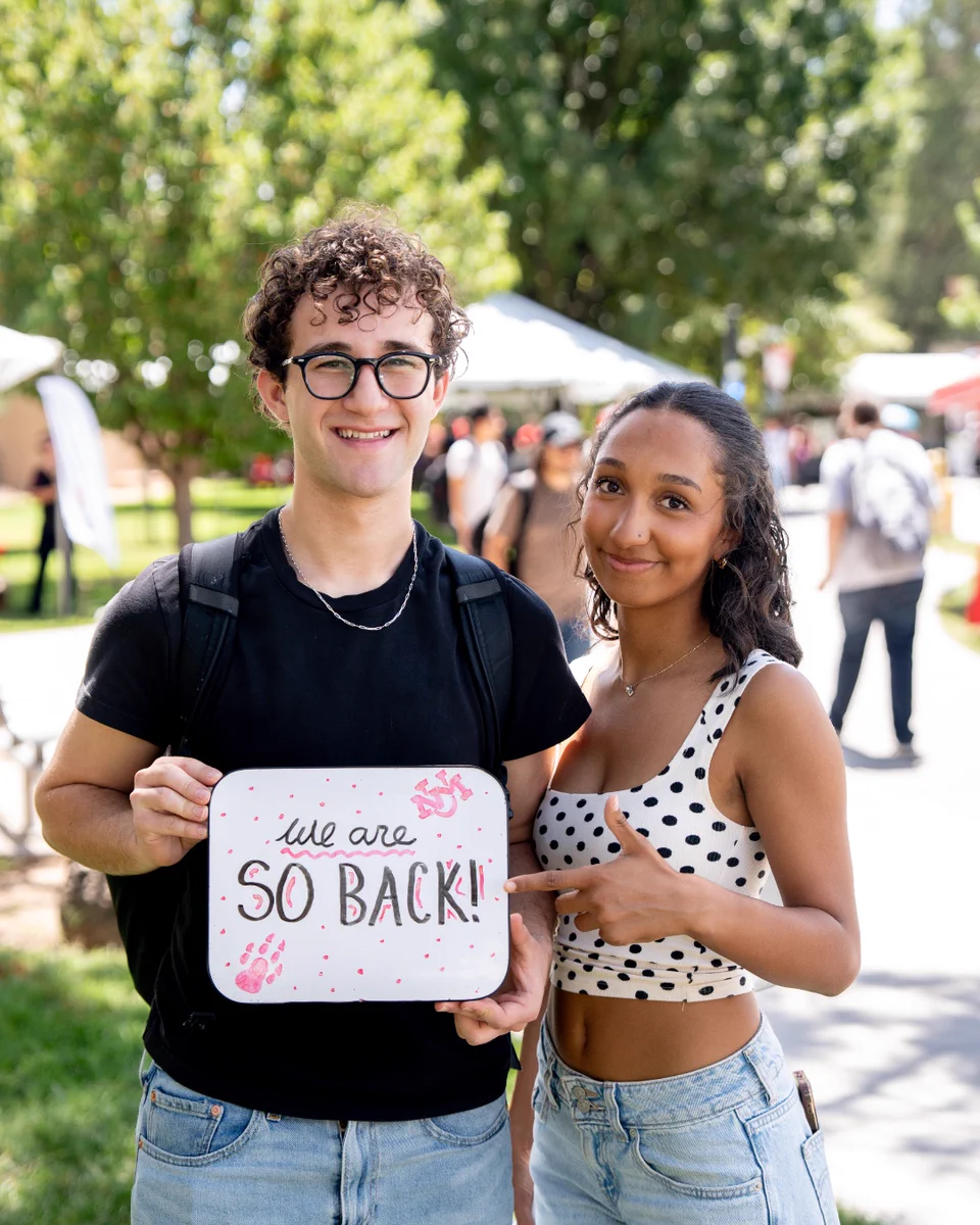 Two people are standing outdoors at UNM main campus, holding a sign that reads "We are so back!" The setting includes trees and people in the background, suggesting a lively, sunny day.