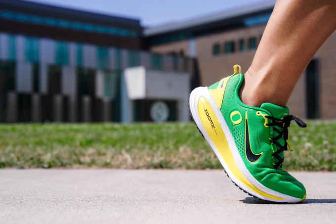A person wearing green Nike Oregon running shoes steps forward on a paved path, with a blurred modern building in the background.