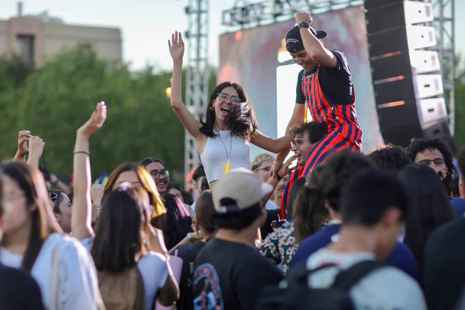 A lively outdoor scene with a cheering crowd at UTEP's Texas Western Gold Rush. One person is smiling, being lifted above others' shoulders, and wearing a striped outfit. Trees and a large screen with colorful lights are visible in the background.
