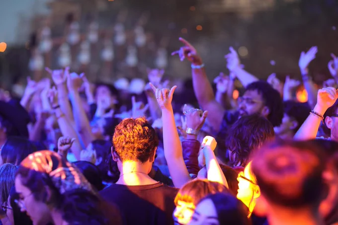 A crowd of people at a concert, with many raising their hands doing UTEP's "picks up" gesture and enjoying the music. The scene is colorful, with concert lighting casting a vibrant glow over the attendees.
