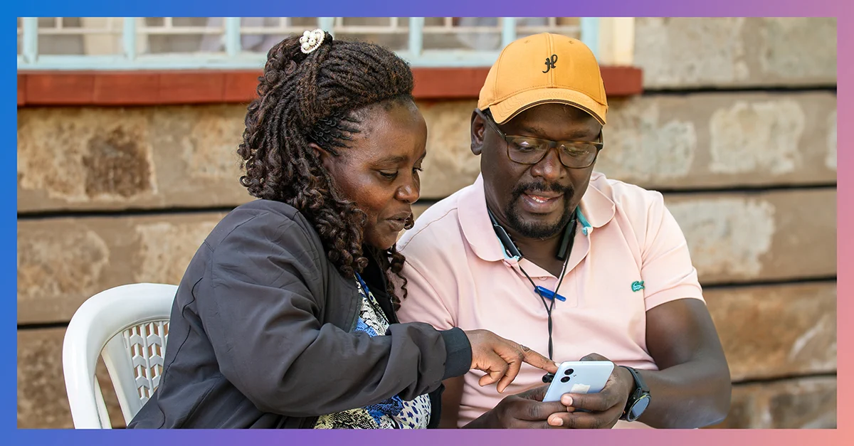 Two people sit side by side outside a building, examining information on a smartphone. One person gestures toward the screen as they review the content together.