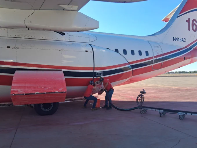Two workers in red shirts refuel a small aircraft on a sunny tarmac under the wing near the rear fuselage.