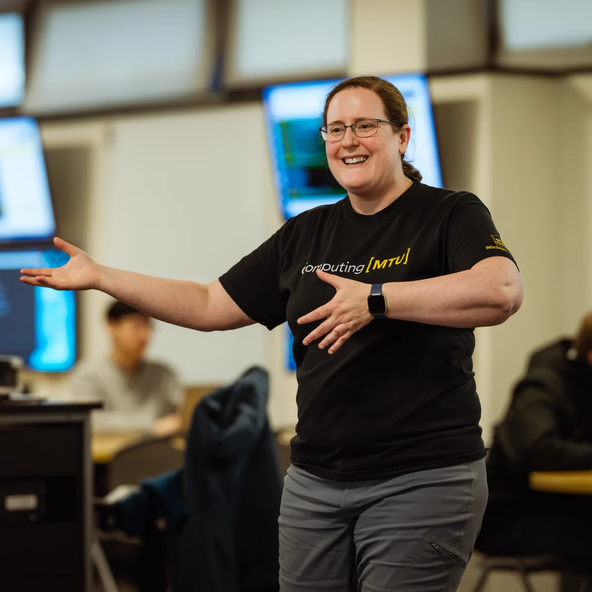 Laura Brown, Chair of the Data Science Department at Michigan Tech, smiles while speaking to students during a lecture. She is wearing glasses, a black Computing MTU shirt, and gray pants.