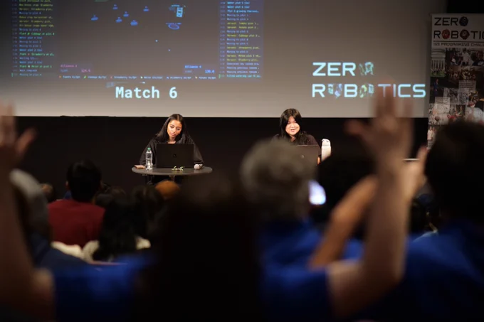 Two young women are seated behind small tables on stage in a darkened auditorium. A large screen behind them displays lines of code and game status text, the words "Match 6," and a "ZERO ROBOTICS" logo. To the right hangs a vertical banner reading "ZERO ROBOTICS ISS Programming Challenge" with a collage of photos. In the foreground, the silhouetted heads and raised hands of audience members are visible.