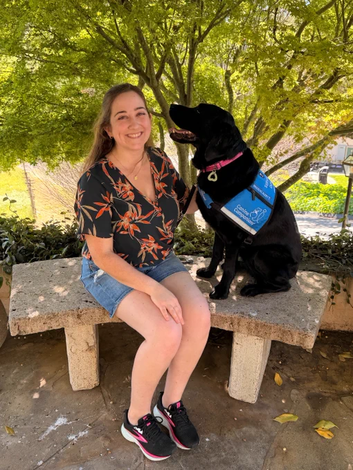 A person is sitting on a stone bench under a tree, smiling at a black dog wearing a blue Canine Companions branded service vest. The scene is outdoors with sunlight filtering through the leaves.