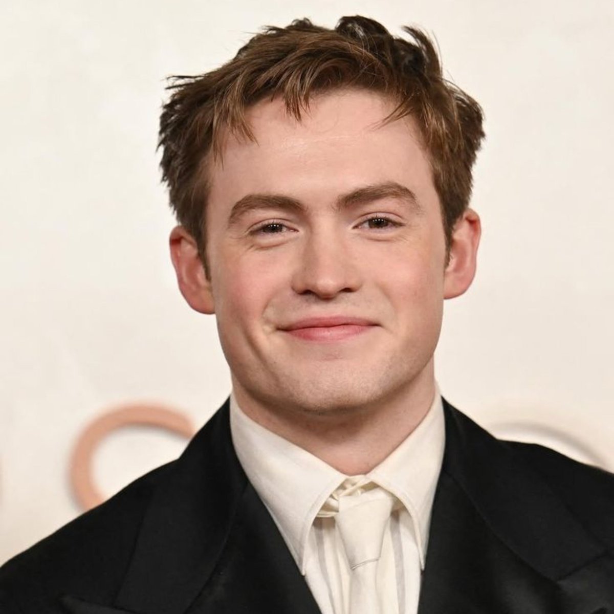 Kit Connor smiles softly in elegant formal wear, light brown hair neatly styled for the red event