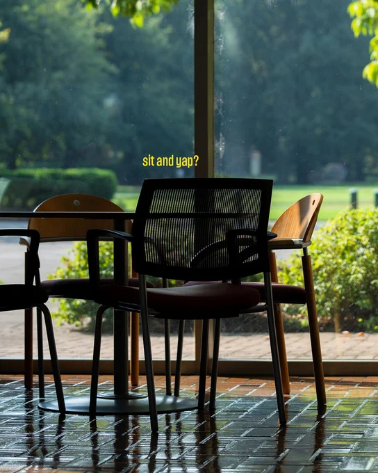 A sunny indoor scene featuring three empty chairs by a large window. The words "sit and yap?" are displayed above the chairs. Outside, a garden with trees and bushes is visible.