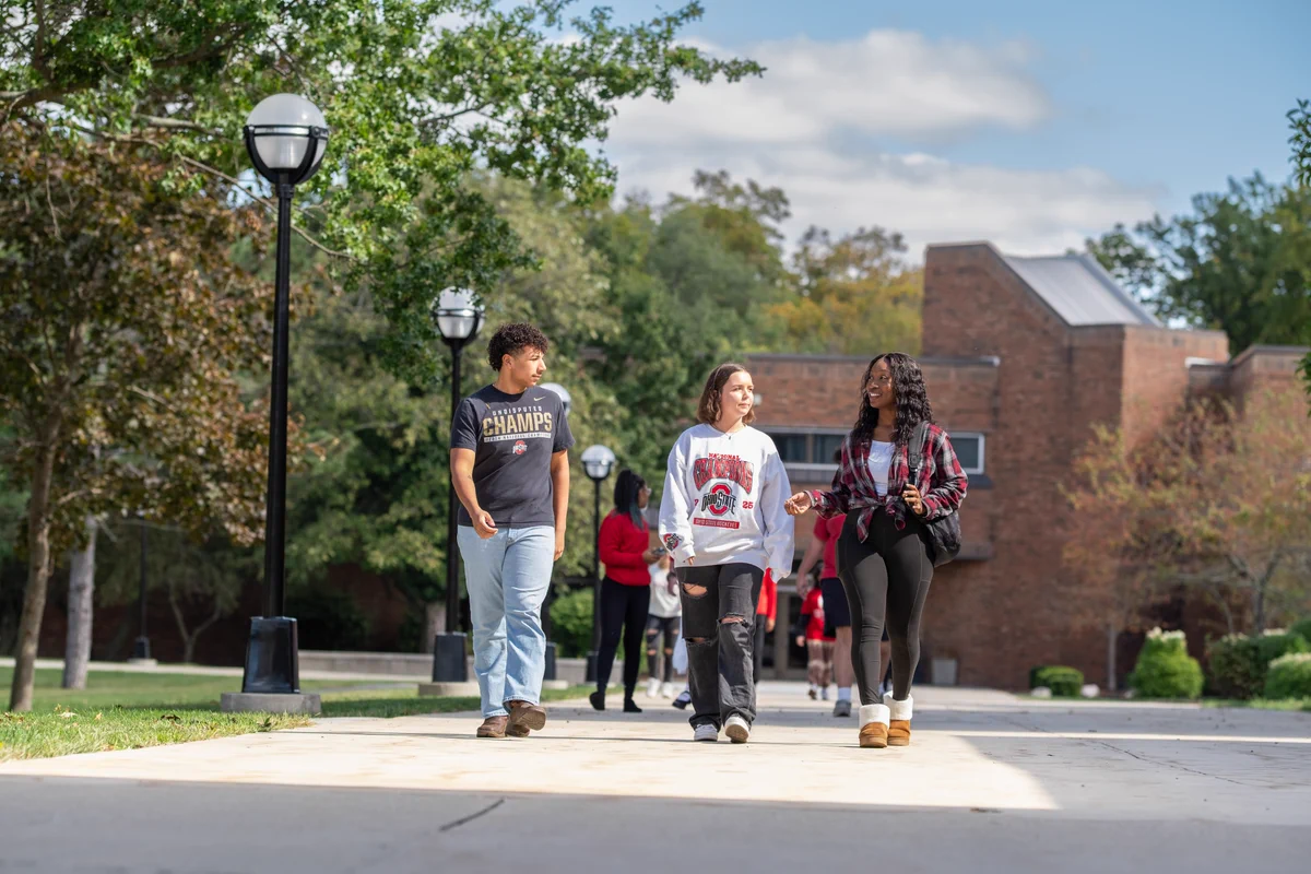 Students walking together on a regional campus pathway with trees and a brick building in the background. They appear to be engaged in conversation and enjoying a sunny day.