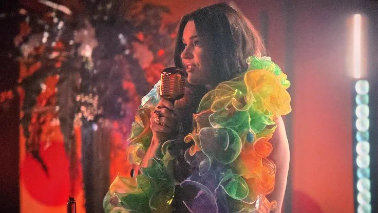 Photo of a TV screen showing a scene from Star Trek: Strange New Worlds. Commander Una Chin-Riley, wearing a voluminous rainbow-colored ruffled boa, sings into a vintage-style microphone on a karaoke stage bathed in warm orange and red light, with colorful floral decorations blurred in the background.