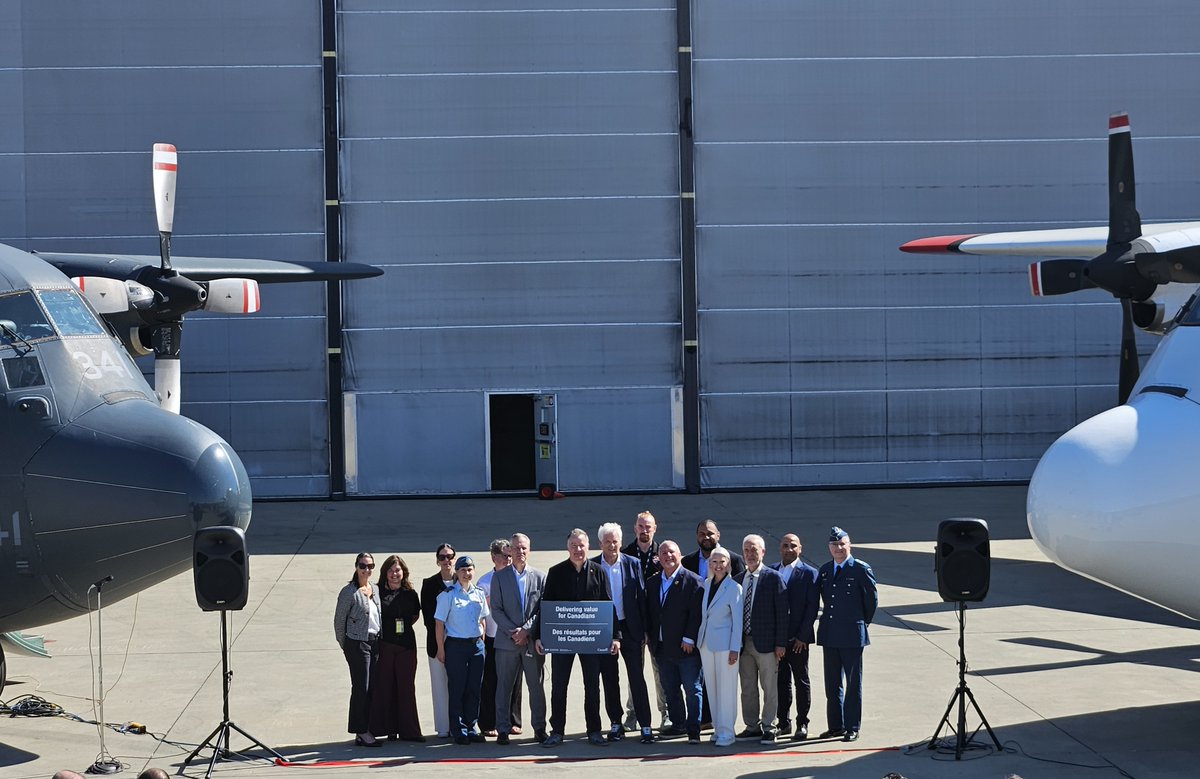 Twelve individuals gathered outdoors on a runway, flanked by twin-engine planes and speakers.